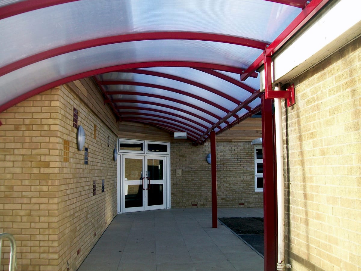 Curved covered walkway in red powder-coated steel with a polycarbonate roof